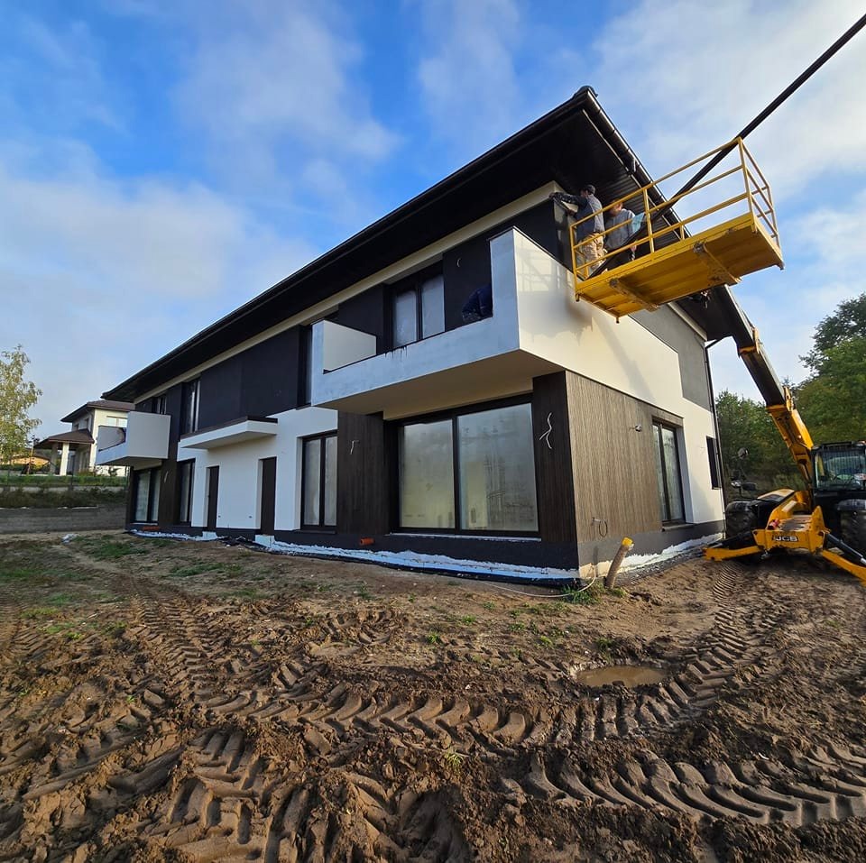 Dwukondygnacyjny dom z ciemną elewacją w trakcie wykańczania; pracownicy na podnośniku koszowym montują elementy wykończeniowe pod dachem.