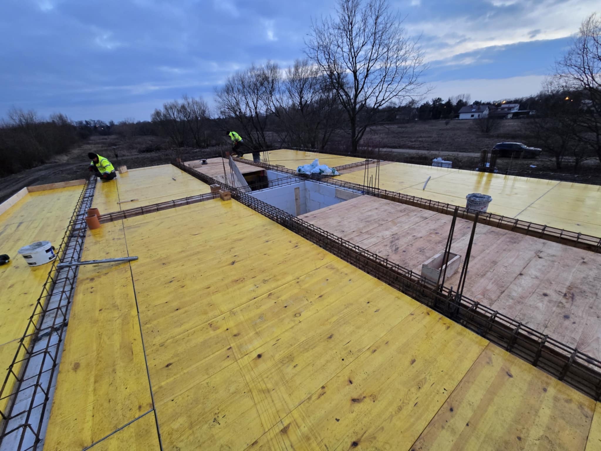Zbrojenie fundamentów domu jednorodzinnego z żółtym szalunkiem, widoczni pracownicy w odblaskowych kamizelkach, w tle zabudowa jednorodzinna i drzewa.