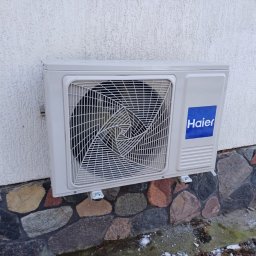 PM GROUP SPÓŁKA Z OGRANICZONĄ ODPOWIEDZIALNOŚCIĄ - Zewnętrzna jednostka klimatyzatora Haier zamontowana na białej ścianie z kamienną podmurówką, widoczne ślady śniegu.