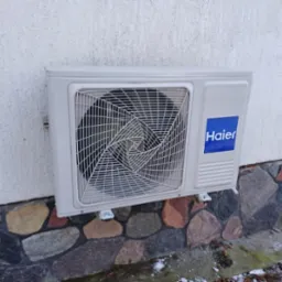 Zewnętrzna jednostka klimatyzatora Haier zamontowana na białej ścianie z kamienną podmurówką, widoczne ślady śniegu.