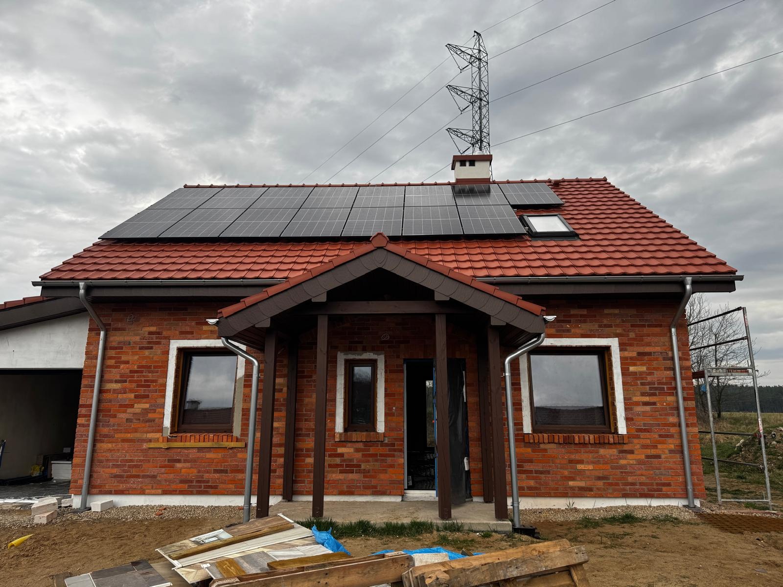 Dom z czerwonej cegły z panelami słonecznymi na dachu z czerwonej dachówki, widoczna linia wysokiego napięcia i rusztowanie z boku.