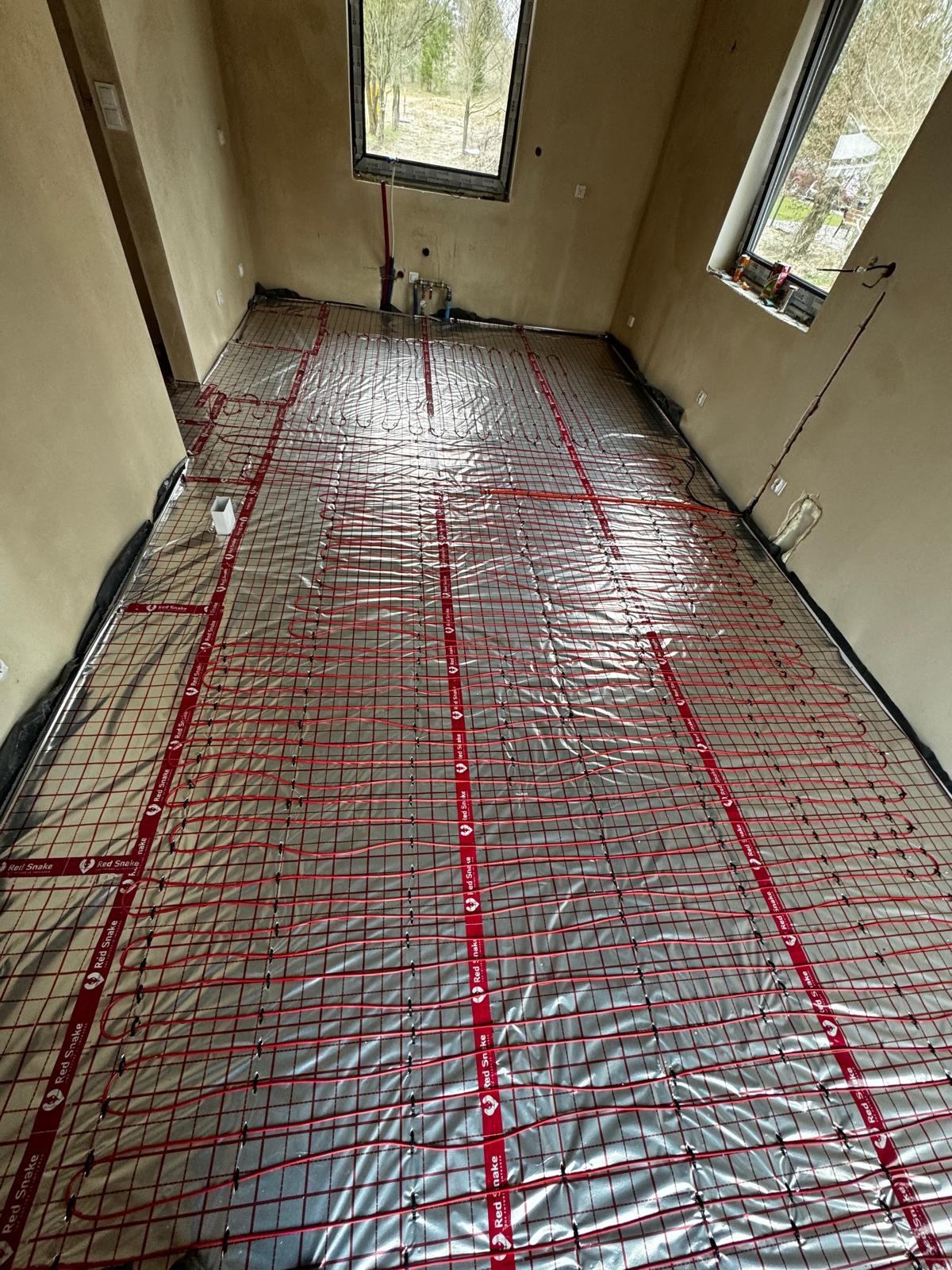 Montaż ogrzewania podłogowego z czerwonymi kablami grzewczymi na folii aluminiowej w pomieszczeniu w Olsztynie, widoczne okna dachowe i instalacja wodna na ścianie.