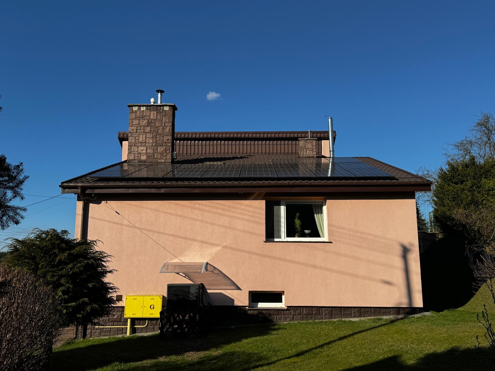 Dom jednorodzinny z brązowym dachem pokrytym panelami fotowoltaicznymi, z ceglanym kominem i żółtą skrzynką gazową na tle zielonego trawnika i błękitnego nieba.