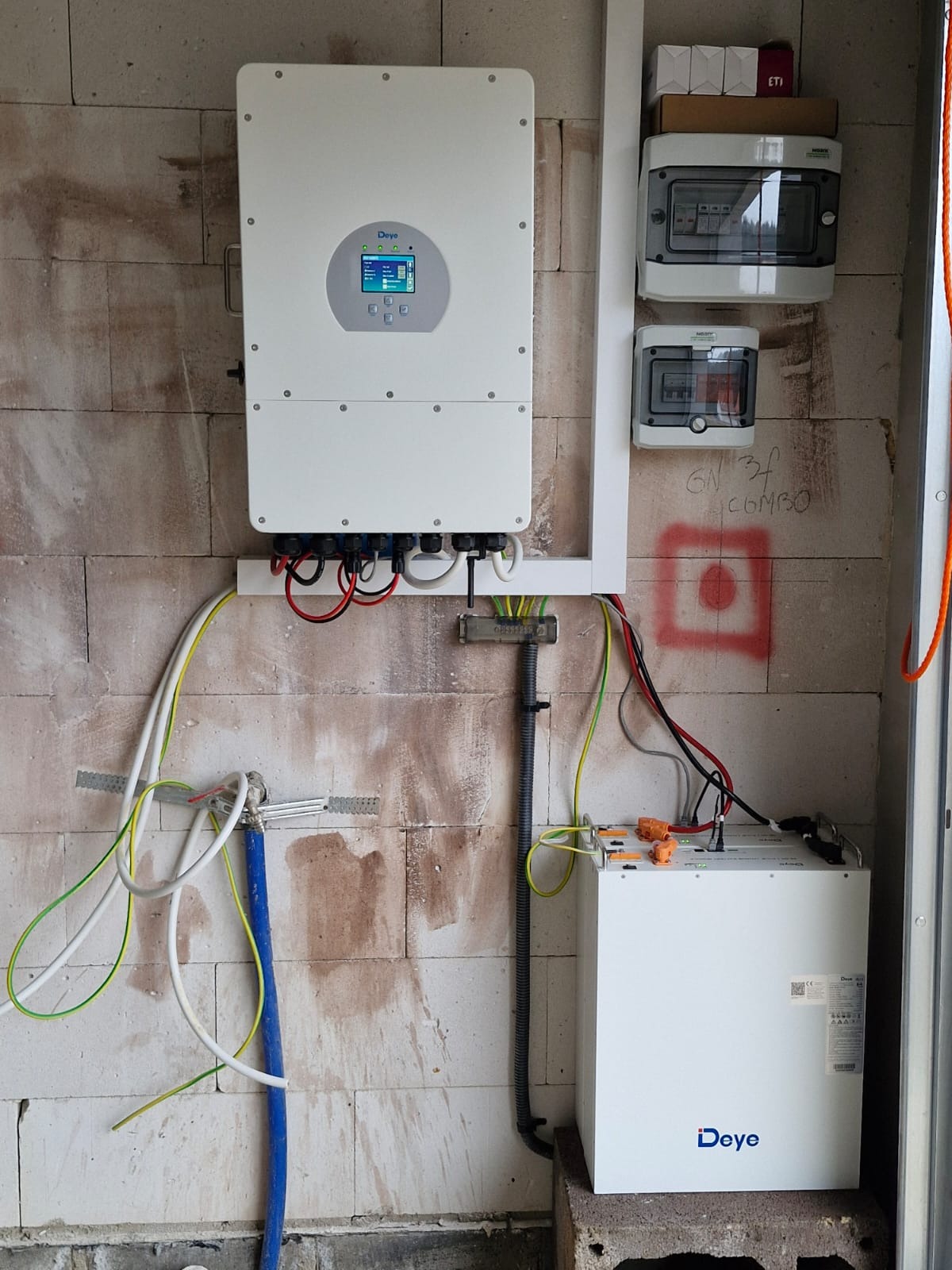 Biały inwerter marki Deye zamontowany na ścianie z betonu komórkowego, z widocznymi kablami, obok skrzynki elektryczne i akumulator magazynu energii Deye na postumencie.