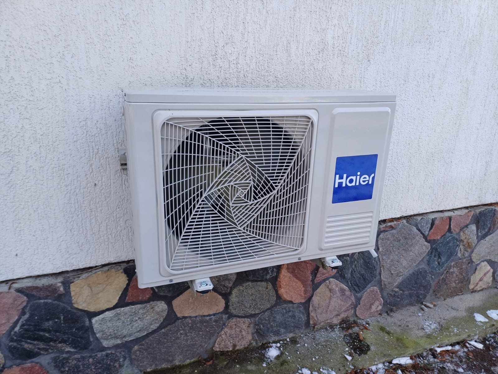 Zewnętrzna jednostka klimatyzatora Haier zamontowana na białej ścianie z kamienną podmurówką, widoczne ślady śniegu.