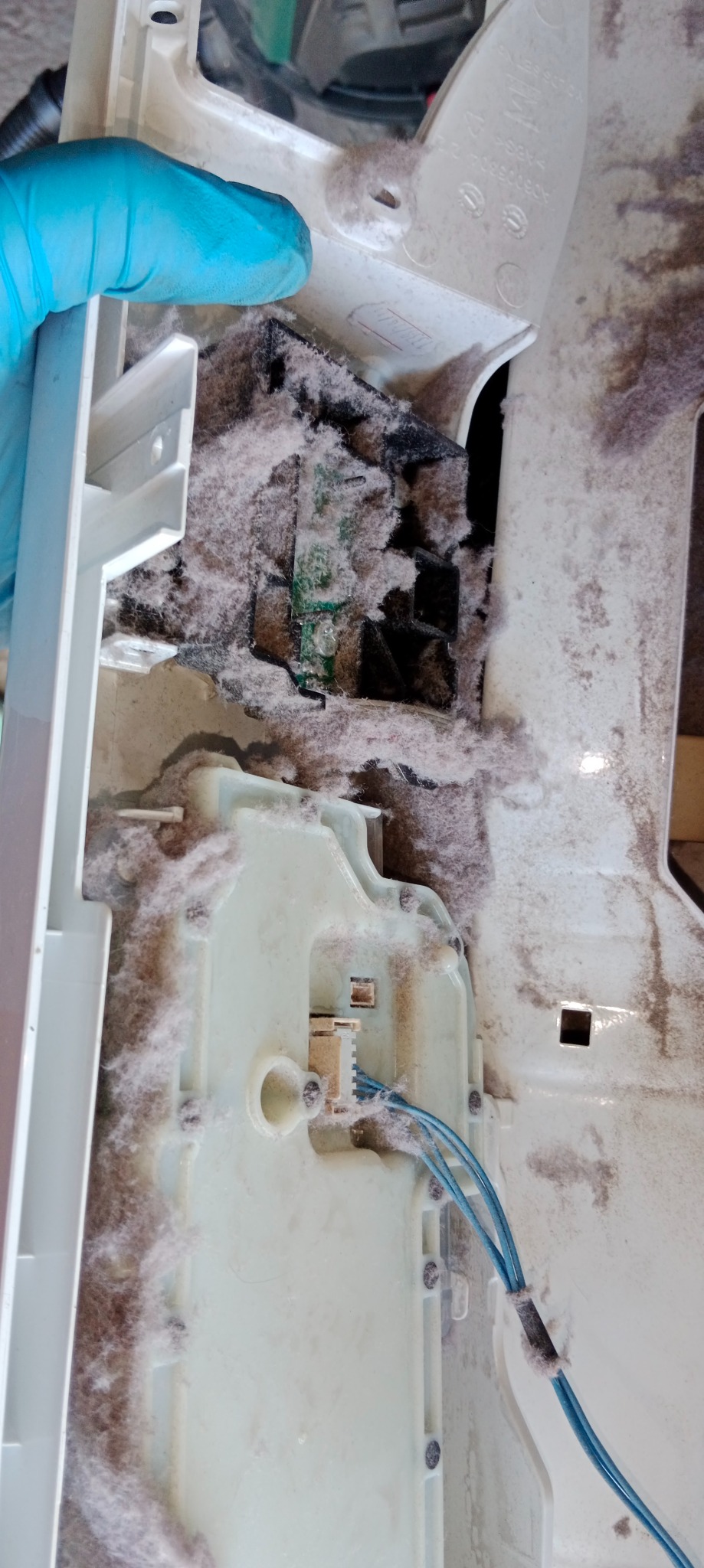 Wnętrze pralki wypełnione kłębami kurzu i włókien, widoczne elementy elektroniczne oraz niebieskie przewody. Serwisant w rękawicy inspekcjonuje wnętrze urządzenia.