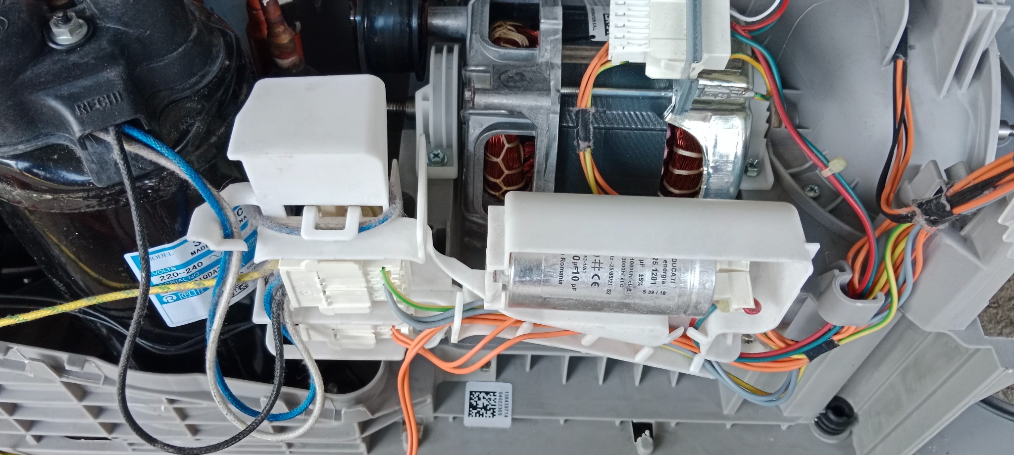 Wnętrze pralki automatycznej: widoczny silnik, kondensator Ducati Energia 25.1281, przewody elektryczne w kolorach pomarańczowym, niebieskim i zielonym oraz czarny kompresor.