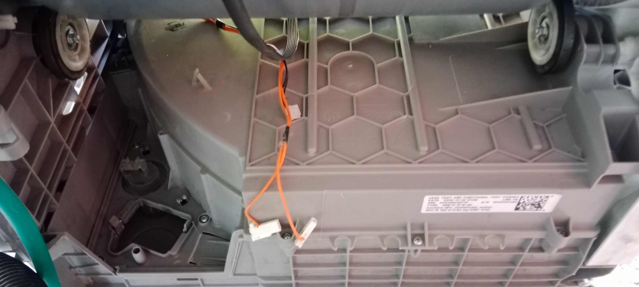 Spód zmywarki z widocznymi elementami konstrukcyjnymi, przewodami i kółkami. Detal z bliska, ukazujący wnętrze urządzenia w trakcie naprawy lub serwisu.