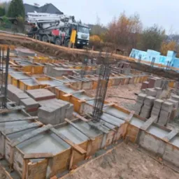 Zalane betonem szalunki fundamentowe z wystającymi prętami zbrojeniowymi, na tle betonomieszarki z pompą i ułożonych bloczków betonowych.