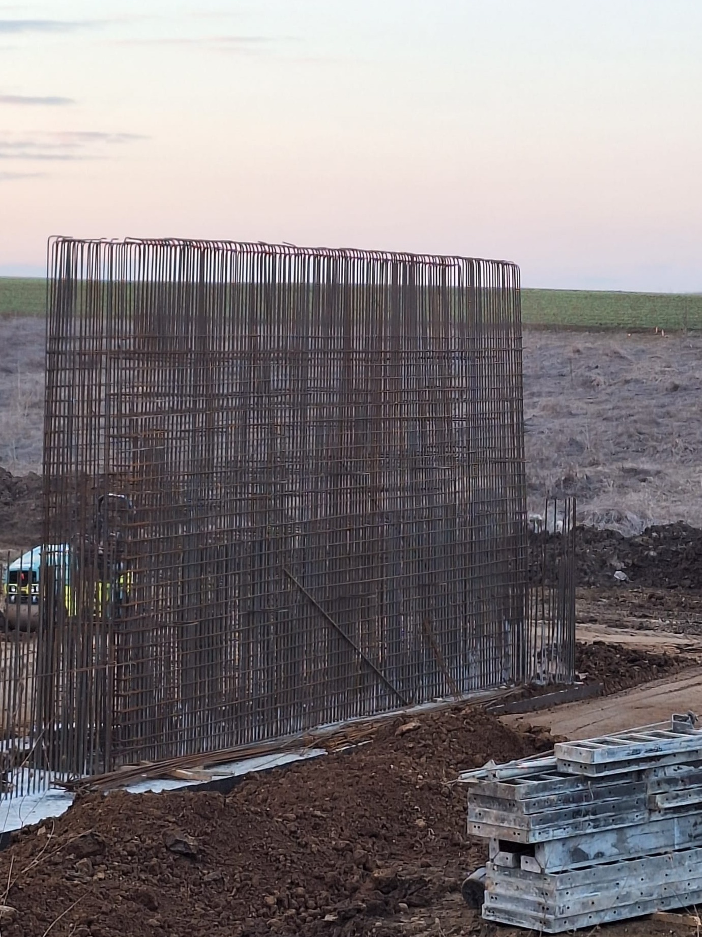 Szkielet z prętów zbrojeniowych na fundamencie, przygotowany do zalania betonem, z widocznymi deskami szalunkowymi w tle i w oddali zieleń pola.