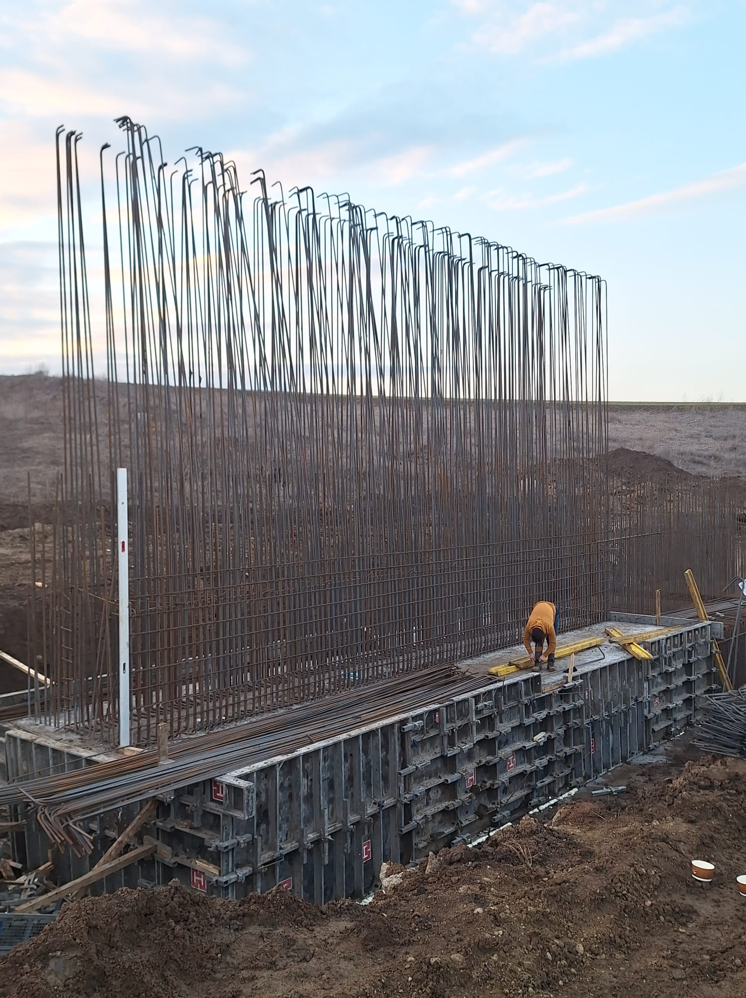 Zbrojenie pionowe i szalunek pod wylewkę fundamentu, pracownik w pomarańczowej bluzie na budowie.