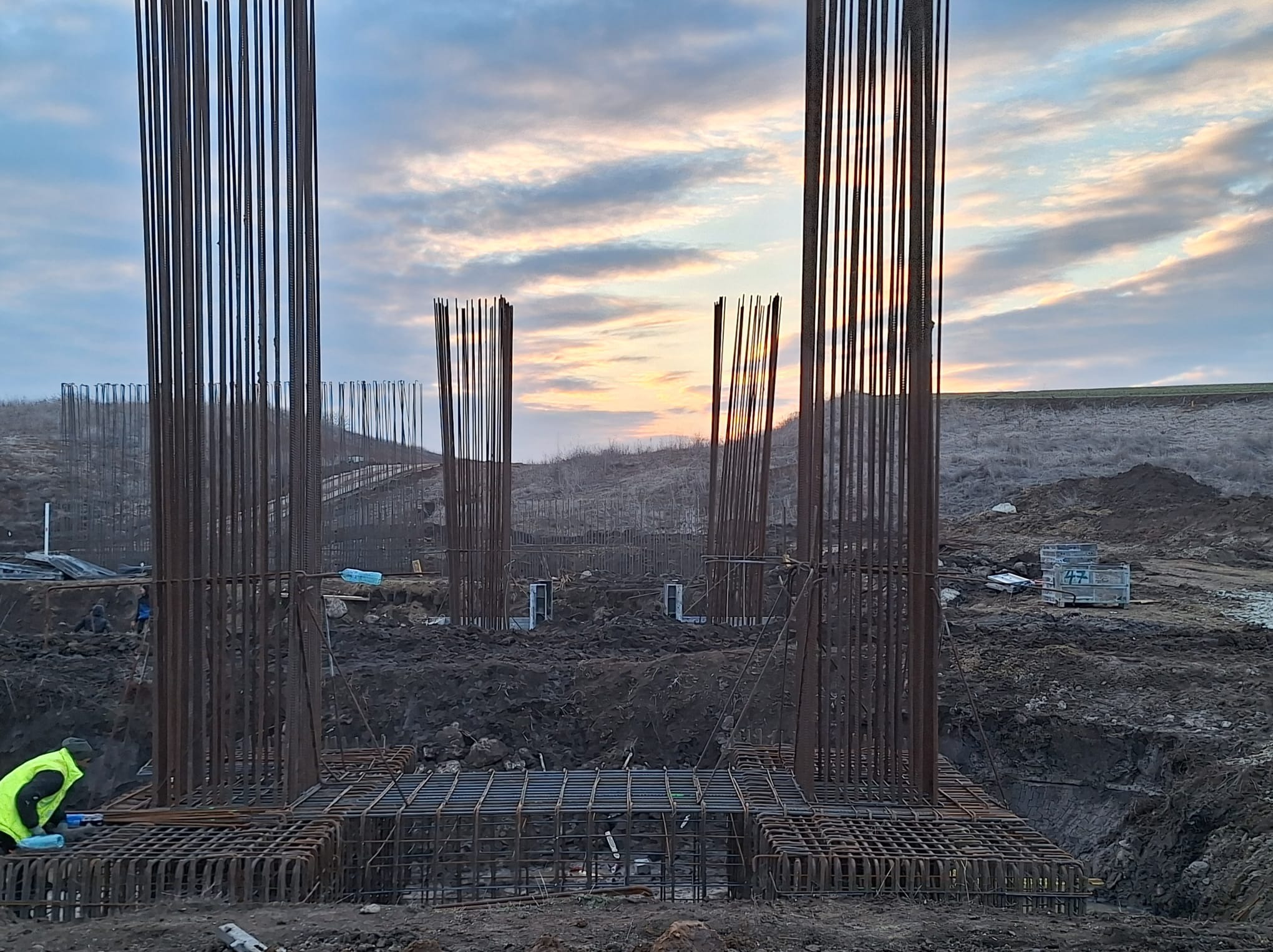 Zbrojenie fundamentów pod budowę, widoczne pionowe pręty i poziome konstrukcje zbrojeniowe, w tle ziemia i zachodzące słońce.