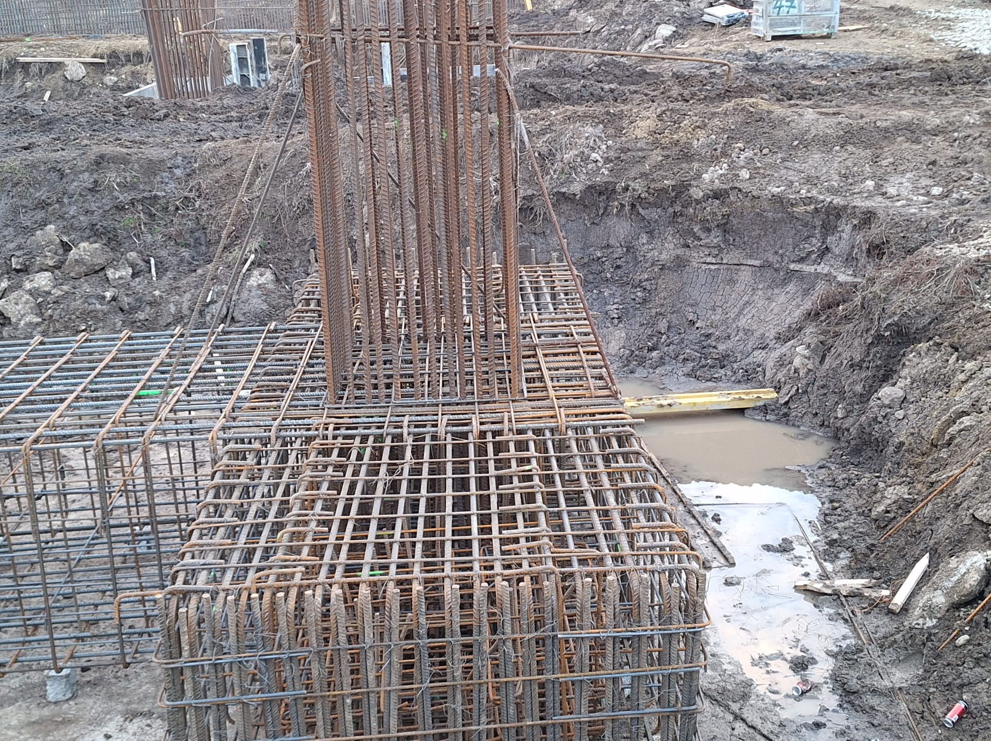 Zbrojenie fundamentu w wykopie wypełnionym błotnistą wodą, widoczne pionowe pręty i siatka zbrojeniowa, teren budowy w stanie surowym.