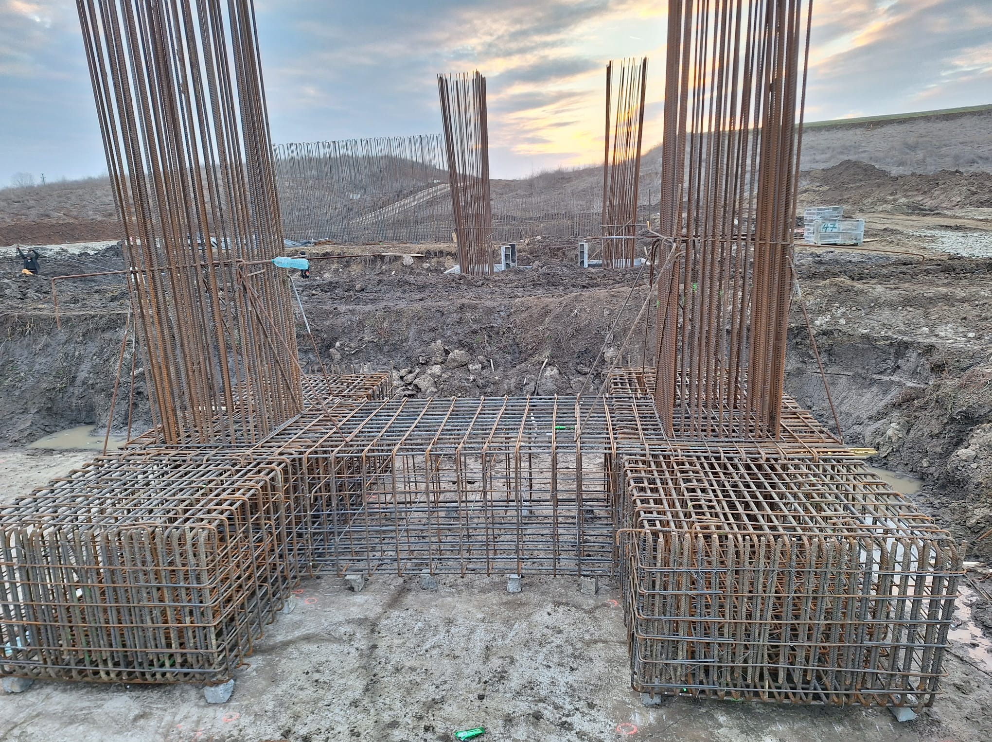 Zbrojenie fundamentów z wystającymi prętami pionowymi na placu budowy, widoczne elementy szalunku w tle, ziemisty teren i zachmurzone niebo.
