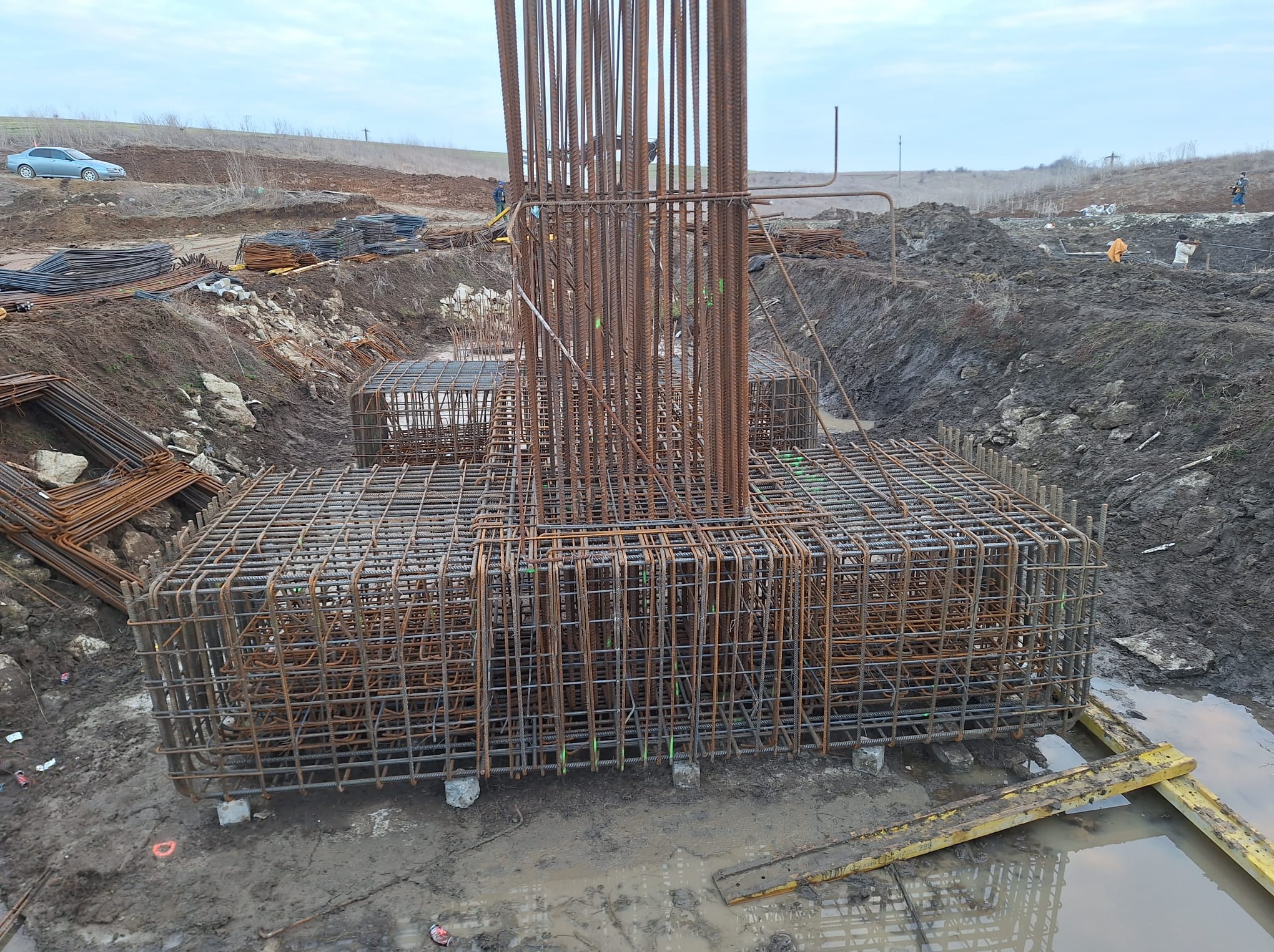 Zbrojenie fundamentu kolumny z wystającymi prętami pionowymi, otoczone błotnistym wykopem, z widocznymi elementami szalunku i zaparkowanym samochodem w tle.
