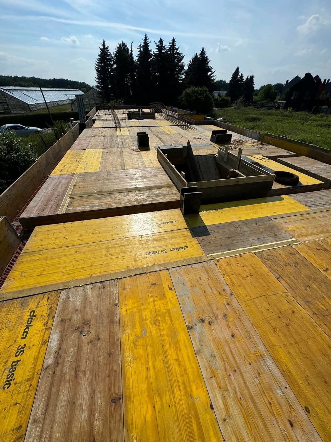 Żółte szalunki Doka 3S basic na deskowaniu fundamentów pod budowę domu, widoczne zbrojenie i otwory technologiczne, w tle zieleń i zabudowania.