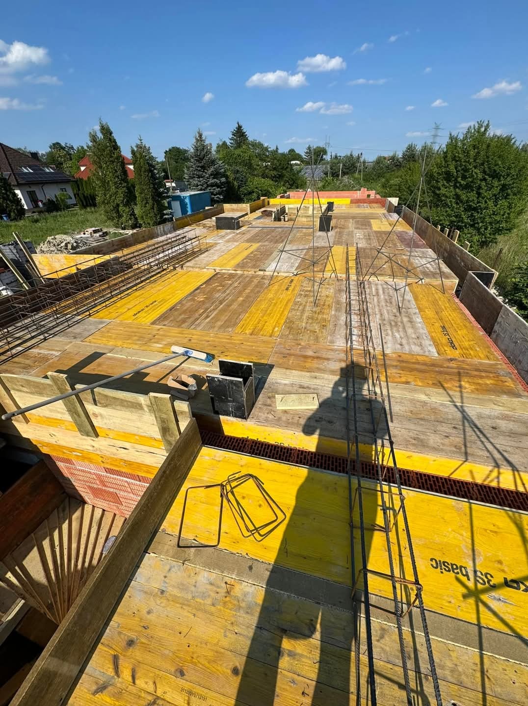 Wiązanie zbrojenia na deskowaniu stropu, widoczne elementy szalunkowe, cegła, narzędzia i cień osoby na budowie domu jednorodzinnego w słoneczny dzień.