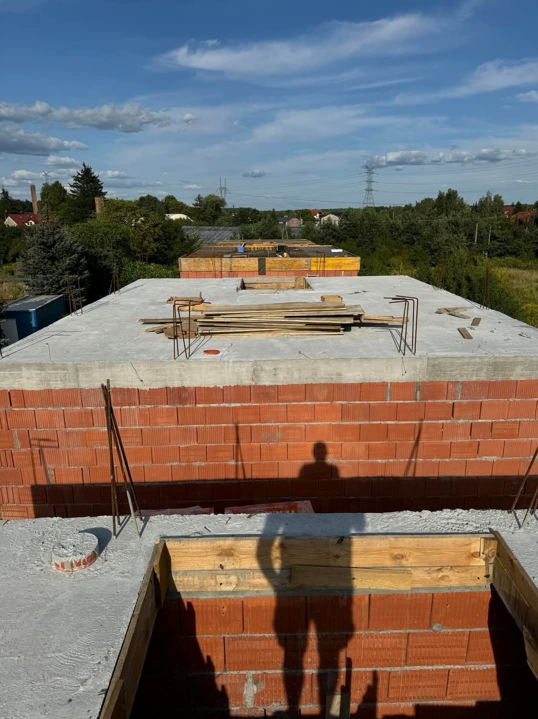 Wznoszenie ścian z czerwonej cegły na betonowym fundamencie, widoczne szalunki i zbrojenia, cień osoby na murze, w tle zabudowa jednorodzinna i błękitne niebo.