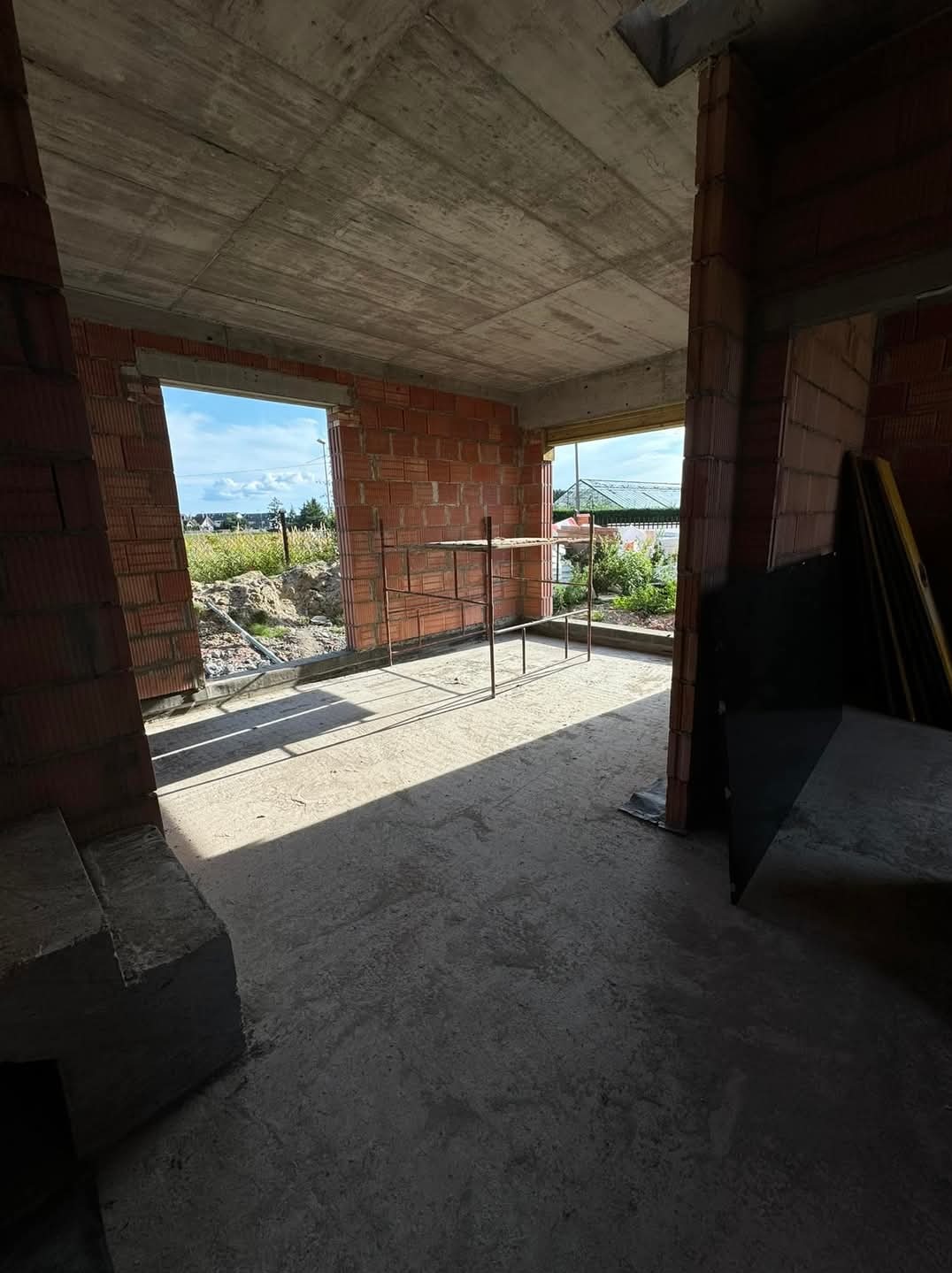 Wnętrze surowego domu w trakcie budowy, widoczne ściany z czerwonej cegły, betonowy strop, metalowe rusztowanie w pokoju i widok na zewnątrz przez niezamknięte otwory okienne.