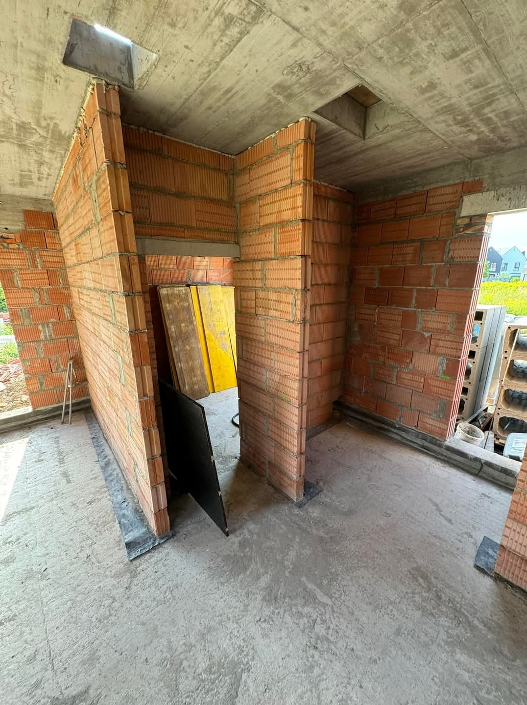 Wnętrze w trakcie budowy z czerwonej cegły ceramicznej, widoczne pionowe ściany działowe, surowy betonowy strop z otworem, żółte płyty szalunkowe oparte o ścianę.