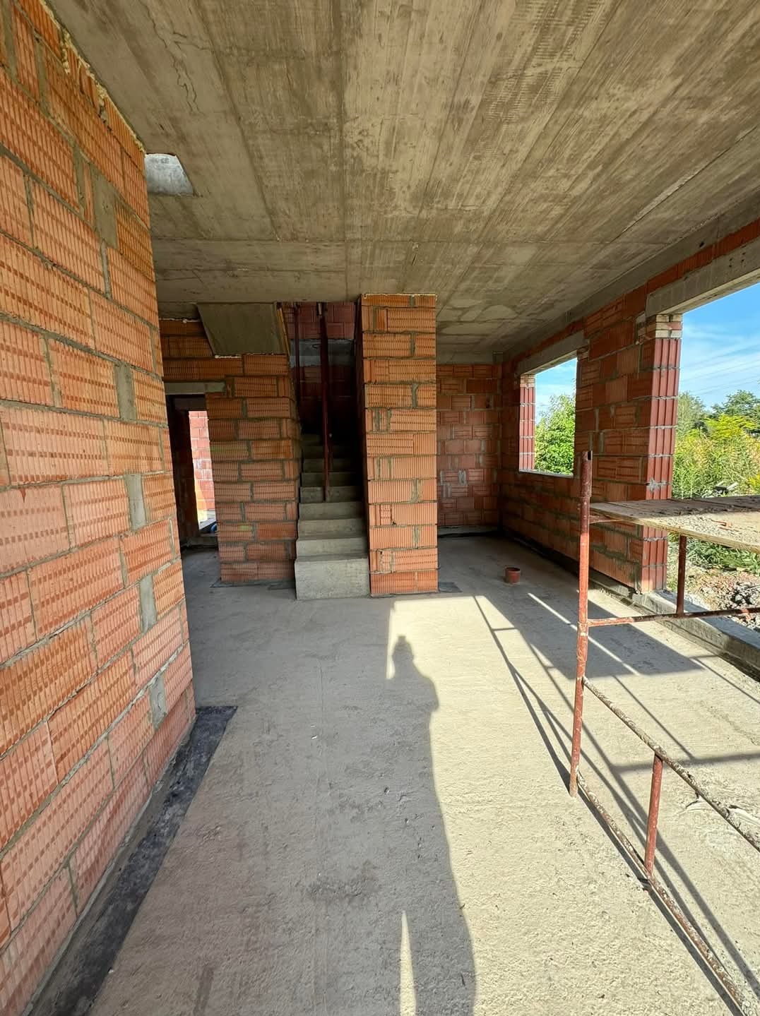 Wnętrze surowego domu w trakcie budowy z cegły ceramicznej, widoczne schody betonowe i niezadaszony balkon z metalowym rusztowaniem, w tle zieleń.