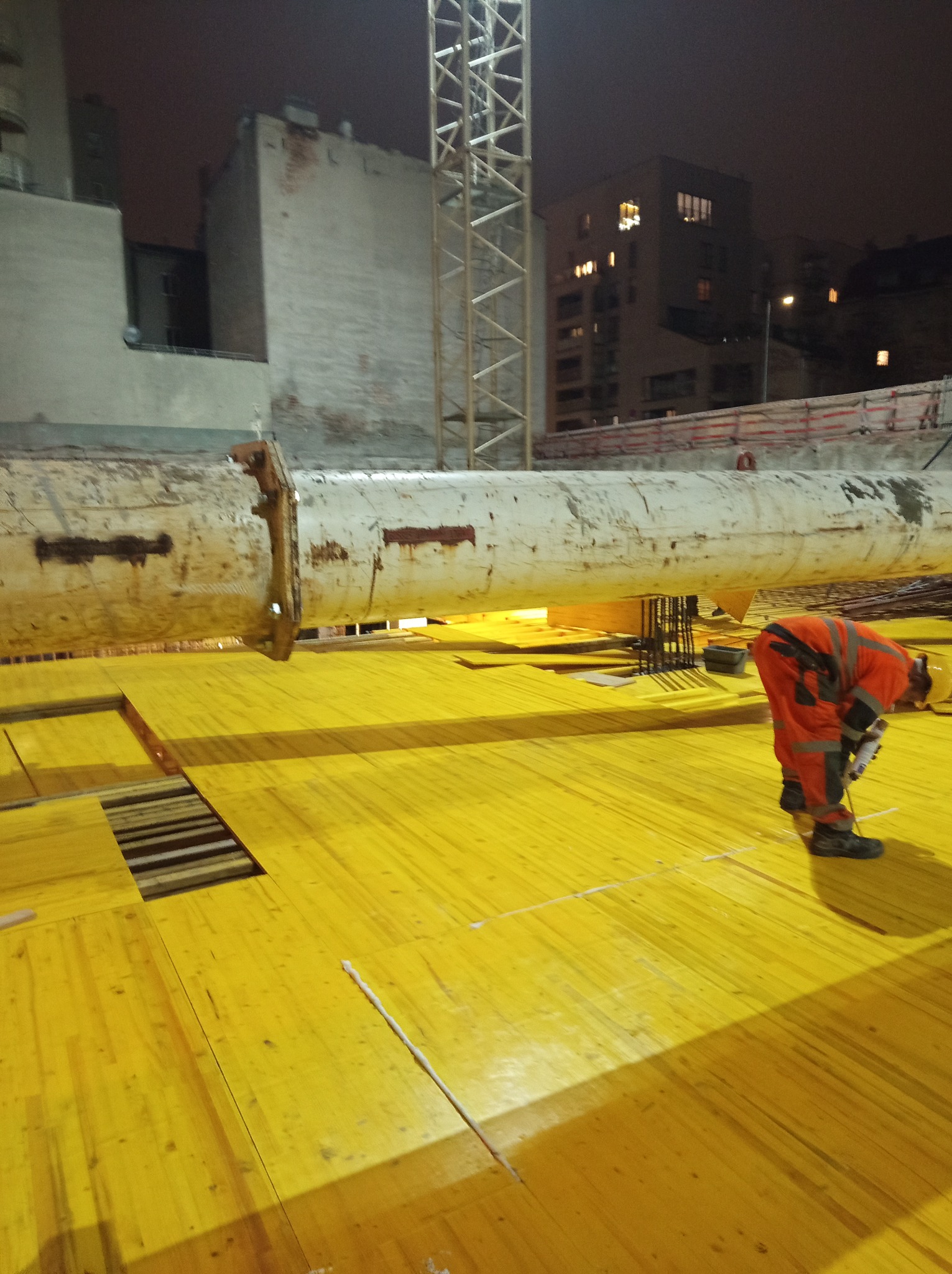 Pracownik w kombinezonie roboczym, pochylony, pracuje na żółtym, drewnianym szalunku podtrzymywanym przez stalowe podpory na placu budowy oświetlonym sztucznym światłem, w tle budynki miejskie...