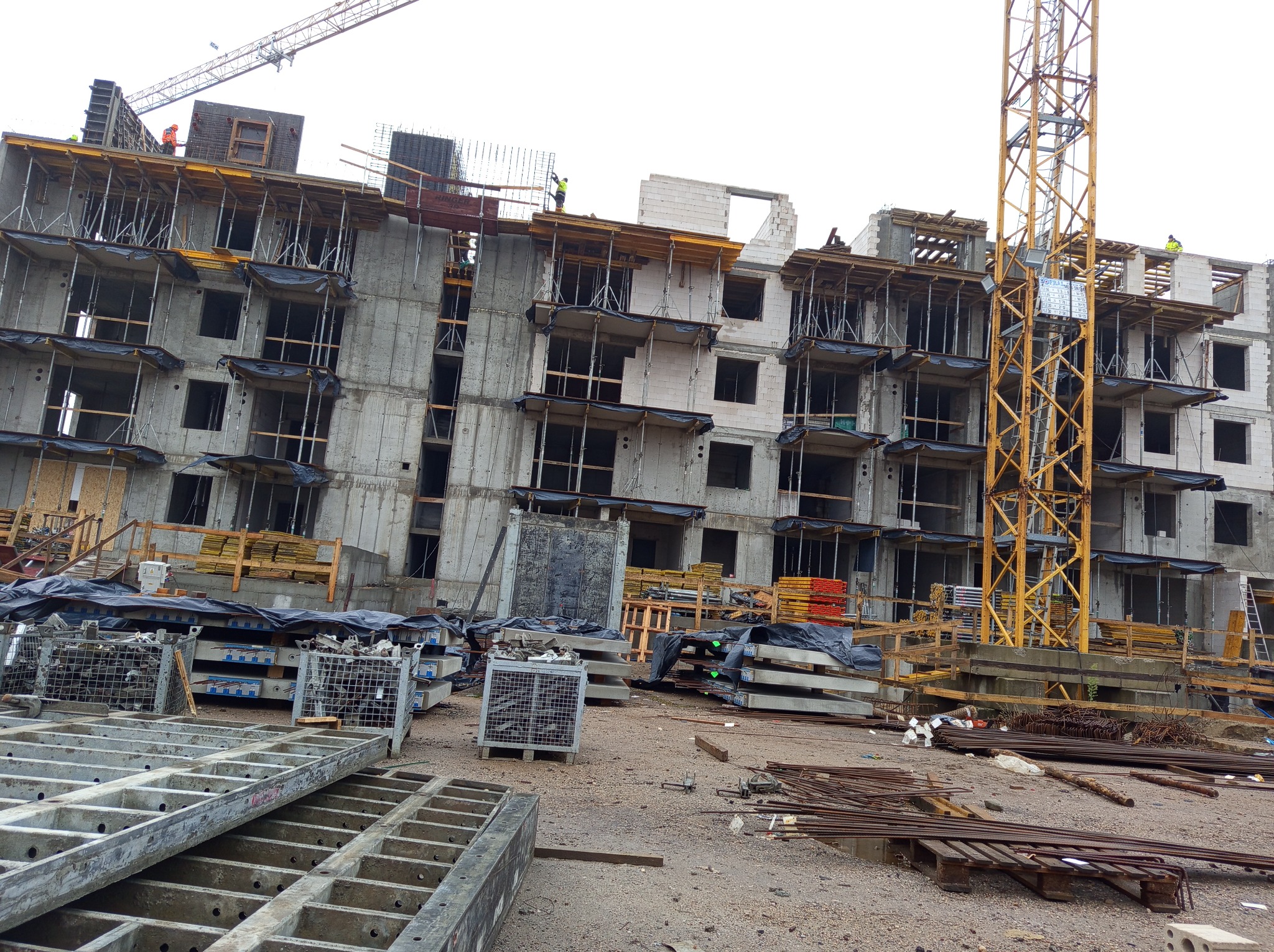 Wielopiętrowy budynek w trakcie wznoszenia ścian z betonu komórkowego, widoczne rusztowania i żuraw budowlany, na pierwszym planie materiały budowlane i elementy konstrukcyjne.