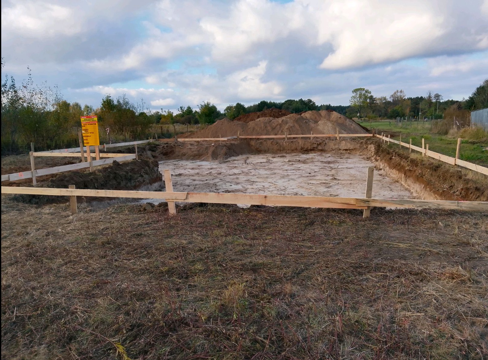 Wykopany fundament pod budowę domu jednorodzinnego, zabezpieczony drewnianymi deskami i stemplami, z widoczną stertą ziemi w tle i tablicą informacyjną.