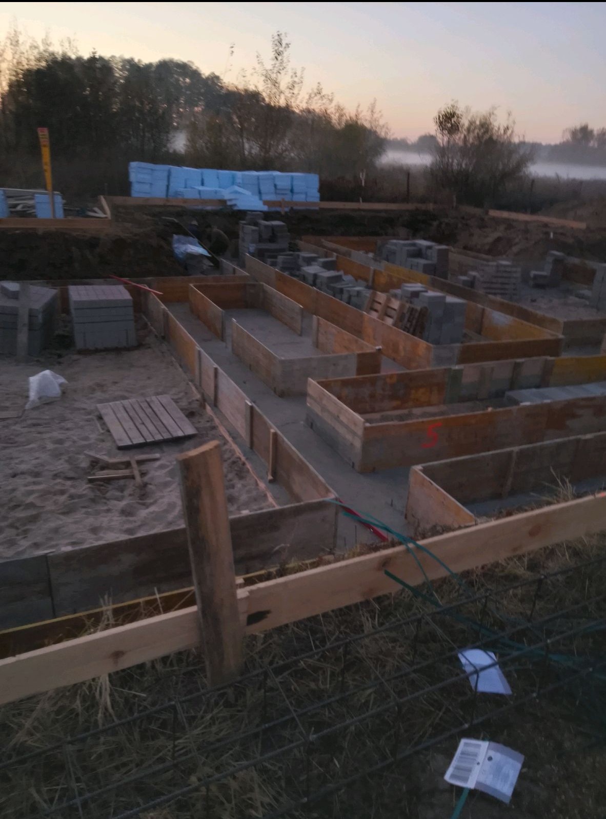 Przygotowanie szalunków pod wylewkę fundamentów domu jednorodzinnego, widoczne bloczki betonowe i stalowe zbrojenie, w tle składowane płyty izolacyjne.