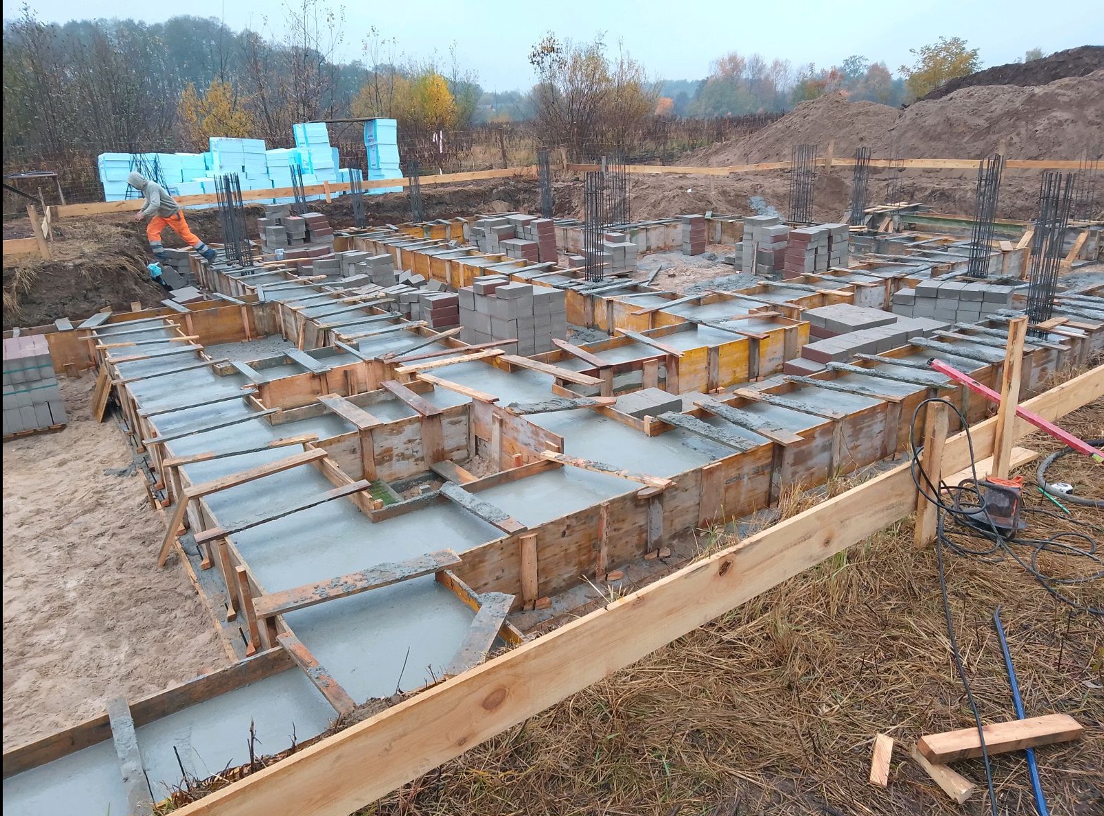 Wylany beton w szalunkach fundamentowych z wystającymi prętami zbrojeniowymi, widoczny pracownik w pomarańczowym kombinezonie oraz zapas bloczków betonowych i styropianu w tle.