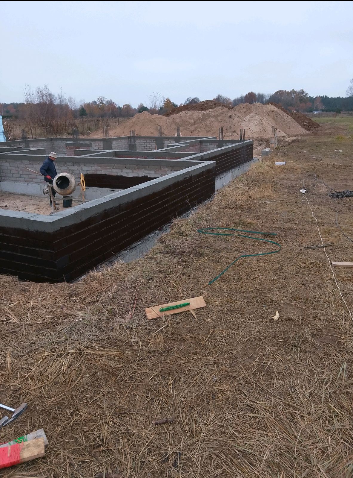 Fundamenty domu jednorodzinnego w Raszynie, z widoczną hydroizolacją ścian fundamentowych, betonową wylewką i pracującym robotnikiem z betoniarką w tle.