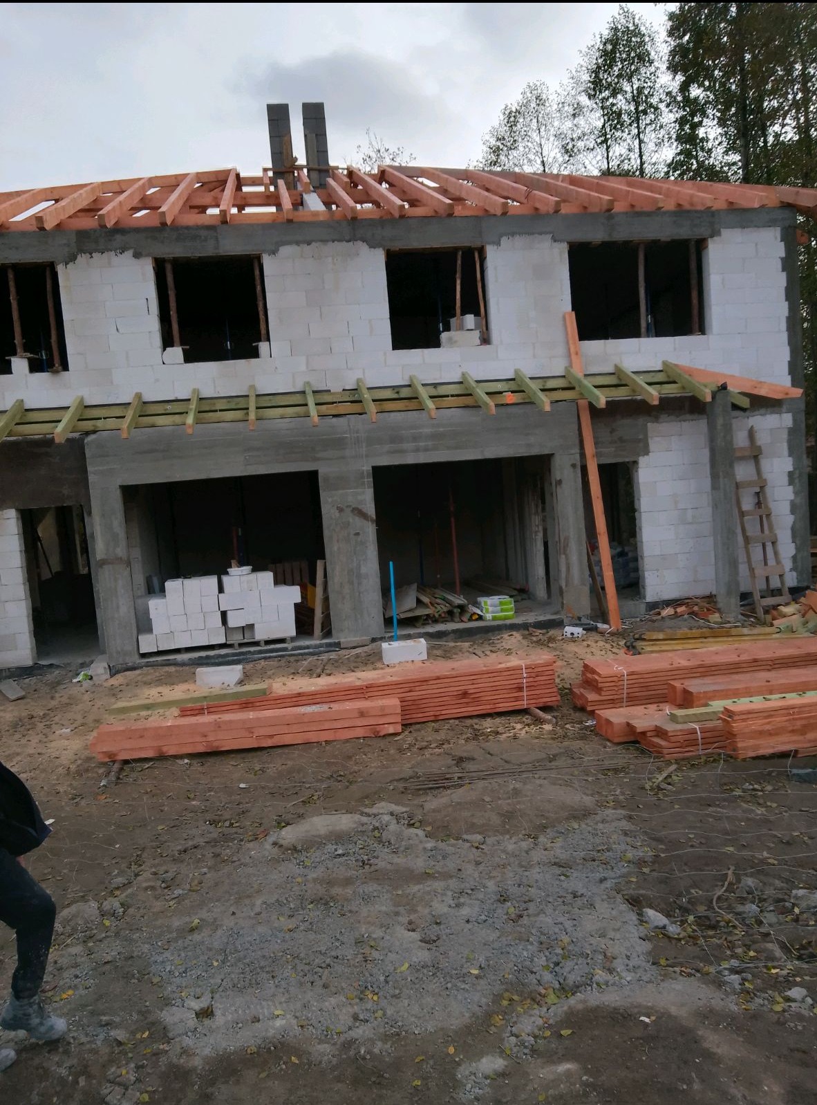 Surowy stan dwupiętrowego domu z białych bloczków, widoczne drewniane elementy konstrukcji dachu i nadproży, palety z deskami na ziemi.