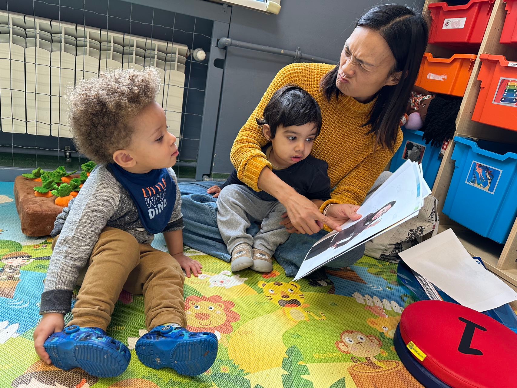 Kobieta w żółtym swetrze czyta książkę dwójce dzieci siedzących na kolorowej macie z motywami zwierzęcymi w pokoju zabaw.