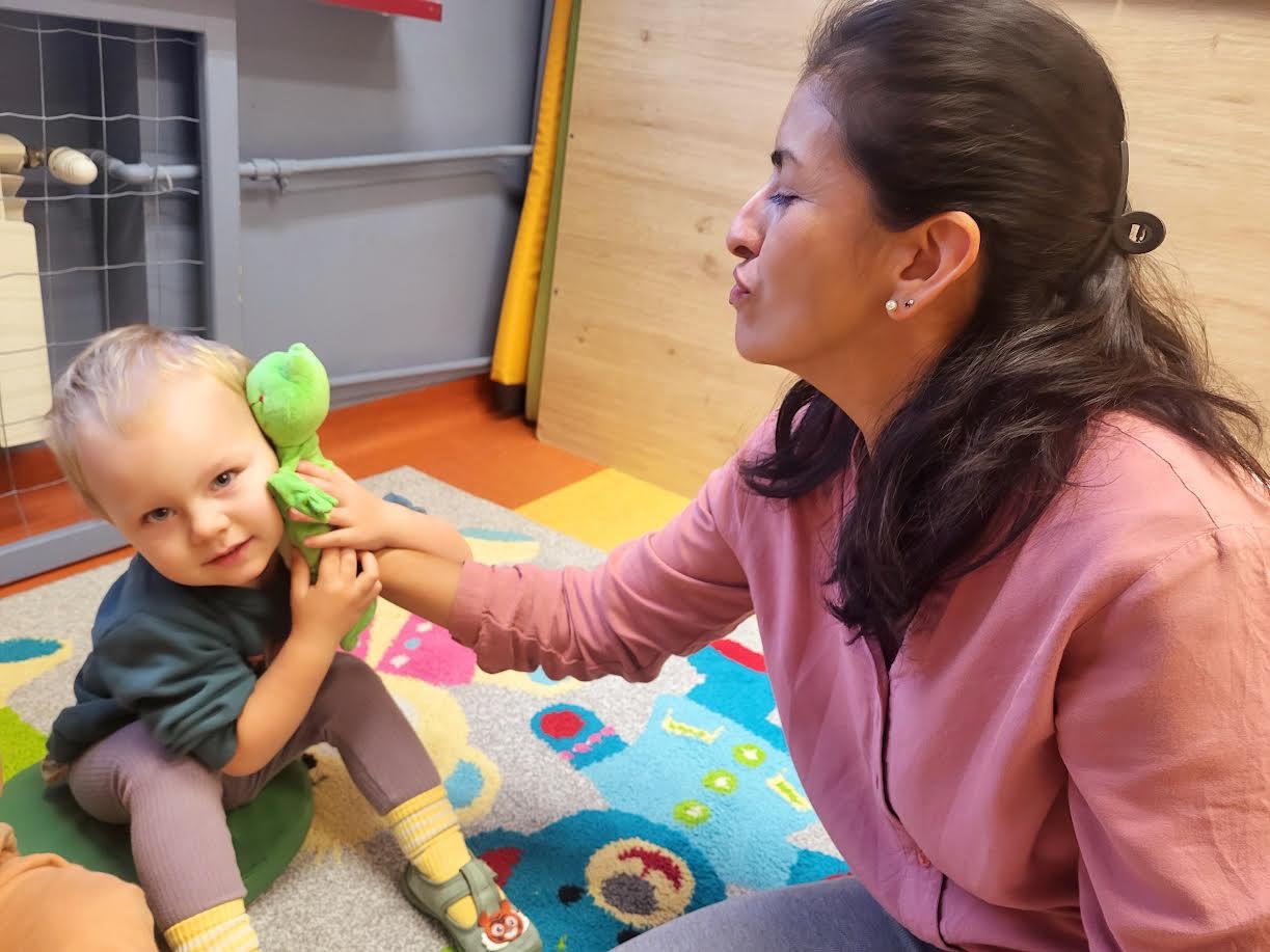 Opiekunka z dzieckiem bawią się pluszową zabawką w żłobku. Kolorowy dywan i jasne otoczenie. Dziecko siedzi na zielonej poduszce, uśmiechając się do kamery.