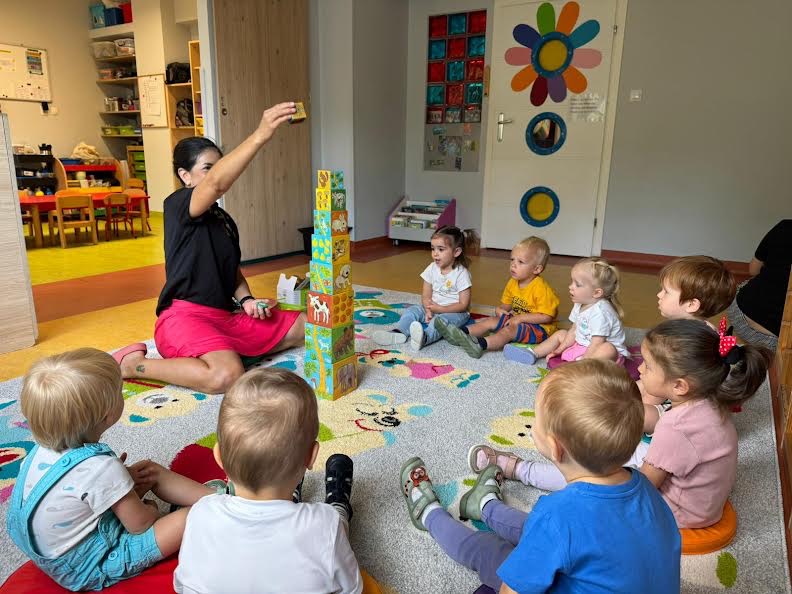 Zajęcia edukacyjne w grupie: opiekunka układa wieżę z klocków z dziećmi siedzącymi na dywanie w jasnym, kolorowym pokoju zabaw.