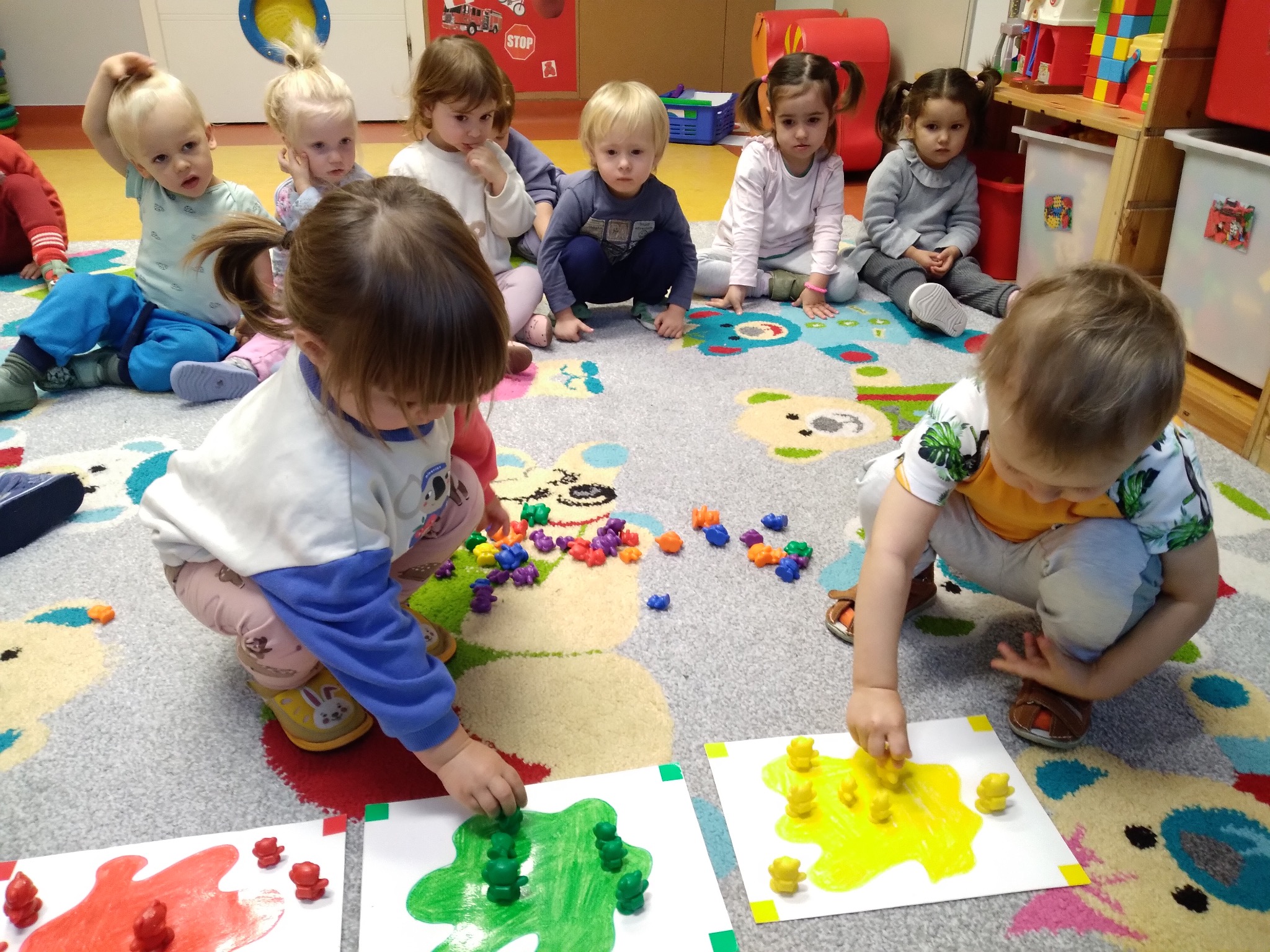 Grupka dzieci w Warszawie bawi się kolorowymi figurkami, układając je na kartkach z namalowanymi plamami w różnych kolorach. Dywan w misie, zabawki w tle.