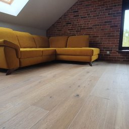 EXOWOOD Kamil Olechowski - Jasne, drewniane deski podłogowe w pomieszczeniu w Żarach z żółtą sofą narożną i ścianą z czerwonej cegły, widok z niskiego kąta.