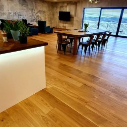 EXOWOOD Kamil Olechowski - Salon z jasną, drewnianą podłogą ułożoną w jodełkę, widok na stół z krzesłami, kanapę i okno z widokiem na zewnątrz.