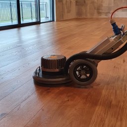 EXOWOOD Kamil Olechowski - Maszyna do renowacji parkietu w trakcie pracy, widoczne świeżo odnowione deski, duże okno balkonowe w tle.