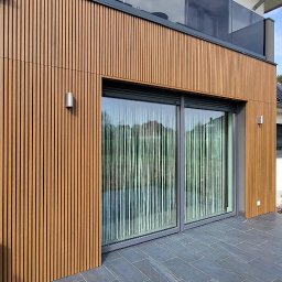 EXOWOOD Kamil Olechowski - Nowoczesna elewacja budynku z drewnopodobnymi panelami pionowymi, dużymi przesuwnymi oknami z widokiem na ogród i tarasem wyłożonym szarymi płytami.