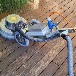 EXOWOOD Kamil Olechowski - Szlifowanie drewnianego tarasu przy użyciu profesjonalnej szlifierki tarczowej z odkurzaczem, widoczne ślady po szlifowaniu i fragment roślinności w tle.