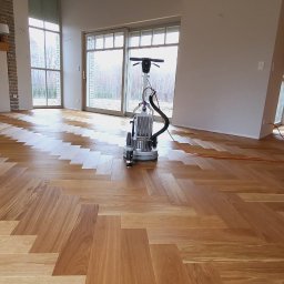 EXOWOOD Kamil Olechowski - Parkiet ułożony w jodełkę podczas cyklinowania maszyną z podłączonym odkurzaczem, widoczne duże okna i fragment ściany z cegły.