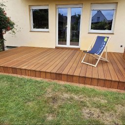 EXOWOOD Kamil Olechowski - Nowoczesny taras z desek, z brązową, ceglaną wędzarnią z boku i krzesłem plażowym z logo, widok z poziomu trawnika na tle domu z oknami.