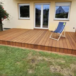 Nowoczesny taras z desek, z brązową, ceglaną wędzarnią z boku i krzesłem plażowym z logo, widok z poziomu trawnika na tle domu z oknami.