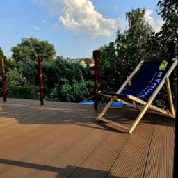 Kompozytowy taras w ciepłym odcieniu brązu z leżakiem z logo firmy, widok na ogród i błękitne niebo z chmurami.