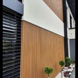 EXOWOOD Kamil Olechowski - Nowoczesna elewacja budynku z pionowymi panelami w kolorze drewna, przedzielona poziomym pasem białego tynku strukturalnego, z czarnymi żaluzjami okiennymi i ozdobnym pasem ziemi z białymi kamykami...