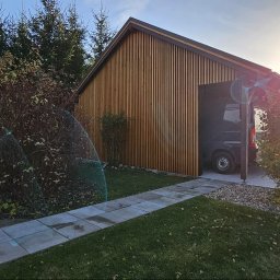 EXOWOOD Kamil Olechowski - Nowoczesna elewacja budynku gospodarczego z pionowymi deskami w odcieniach brązu, widoczny fragment chodnika z płyt betonowych, trawnik i krzewy, w tle zaparkowany samochód dostawczy.