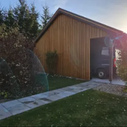 Nowoczesna elewacja budynku gospodarczego z pionowymi deskami w odcieniach brązu, widoczny fragment chodnika z płyt betonowych, trawnik i krzewy, w tle zaparkowany samochód dostawczy.