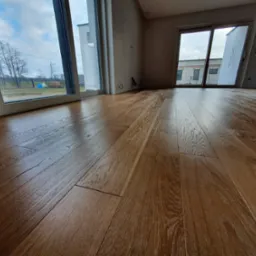 Świeżo ułożona, lakierowana podłoga z desek o naturalnym odcieniu drewna w jasnym wnętrzu z dużymi oknami i widokiem na pochmurne niebo i drzewa.