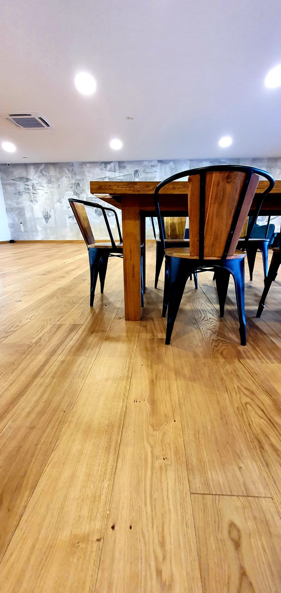 Jasna, drewniana podłoga z widocznymi słojami, uzupełniona o stół z litego drewna i krzesła z czarnymi metalowymi nogami i drewnianym oparciem, na tle ściany z abstrakcyjnym, szarym wzorem.